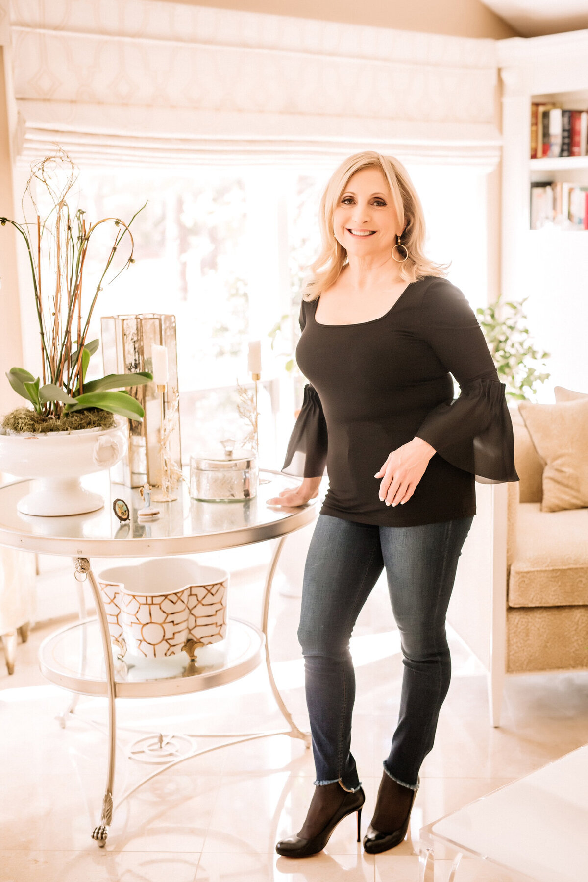 A female dating portrait client standing in her sunroom, leaning against a table, wearing a black blouse & jeans