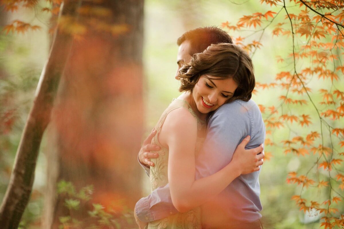 A man embracing a woman from behind, both smiling, amidst a backdrop of soft-focused, autumn-colored leaves.