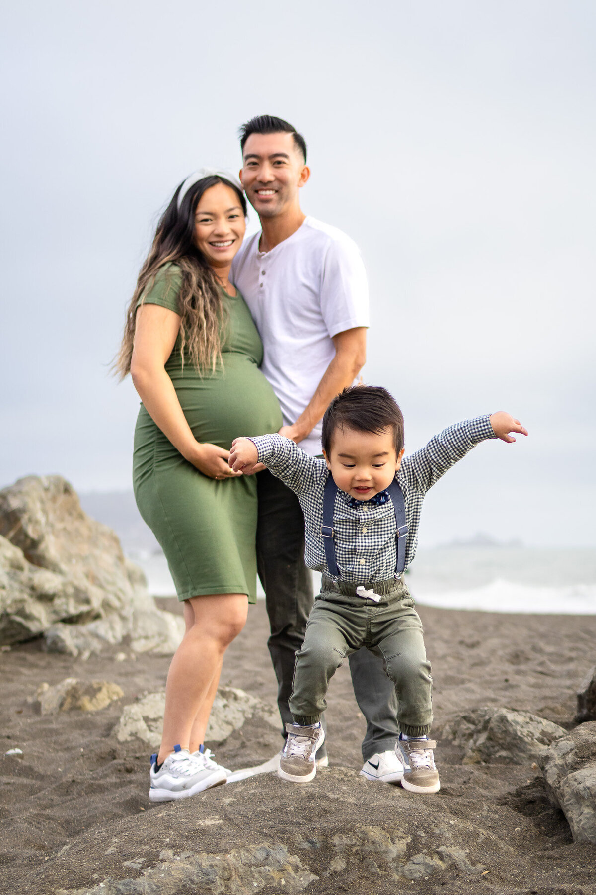 Family maternity photo with parents smiling and toddler jumping in front, featured in Bay Area Maternity Portfolio - Ellobelle Photography