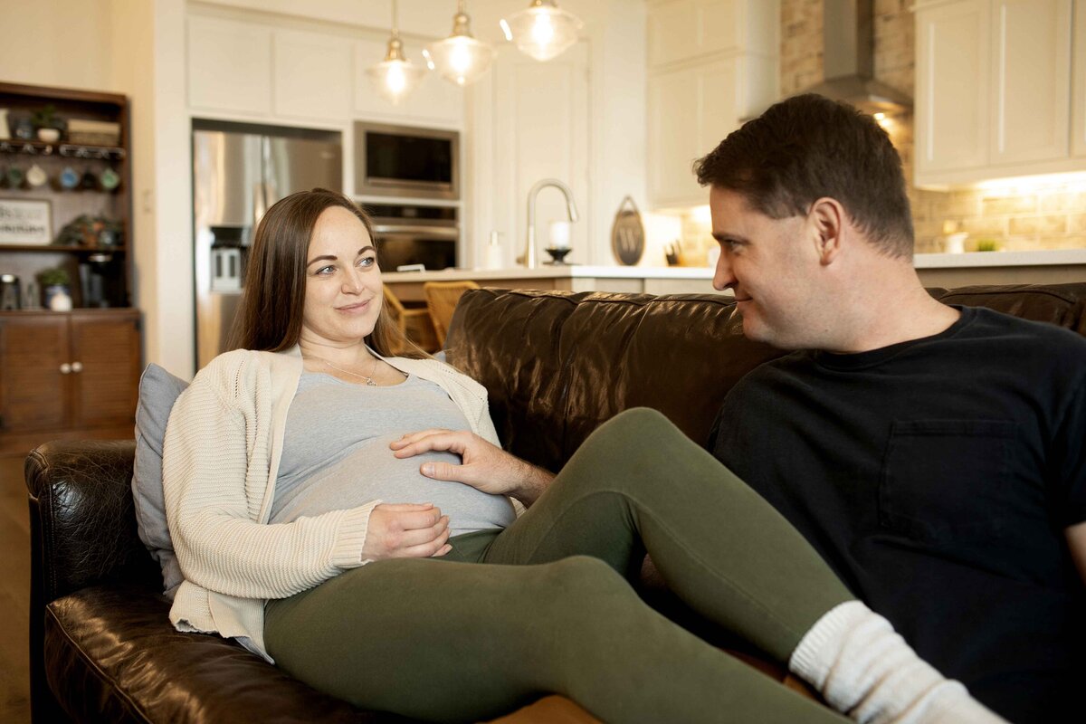 A cozy maternity photo of mom and dad sitting on the couch, with mom cradling her baby bump and dad gently resting his hand on her belly, both sharing a peaceful and loving moment, captured by Chunky Monkey Photography.