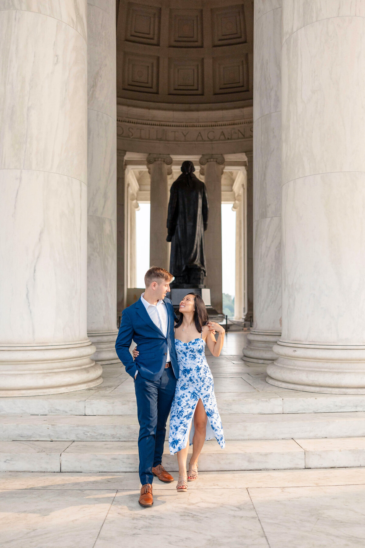 A couple embraces on the steps of a historical monument featuring tall columns and a large statue in the background. The man wears a blue suit, and the woman wears a white and blue floral dress.