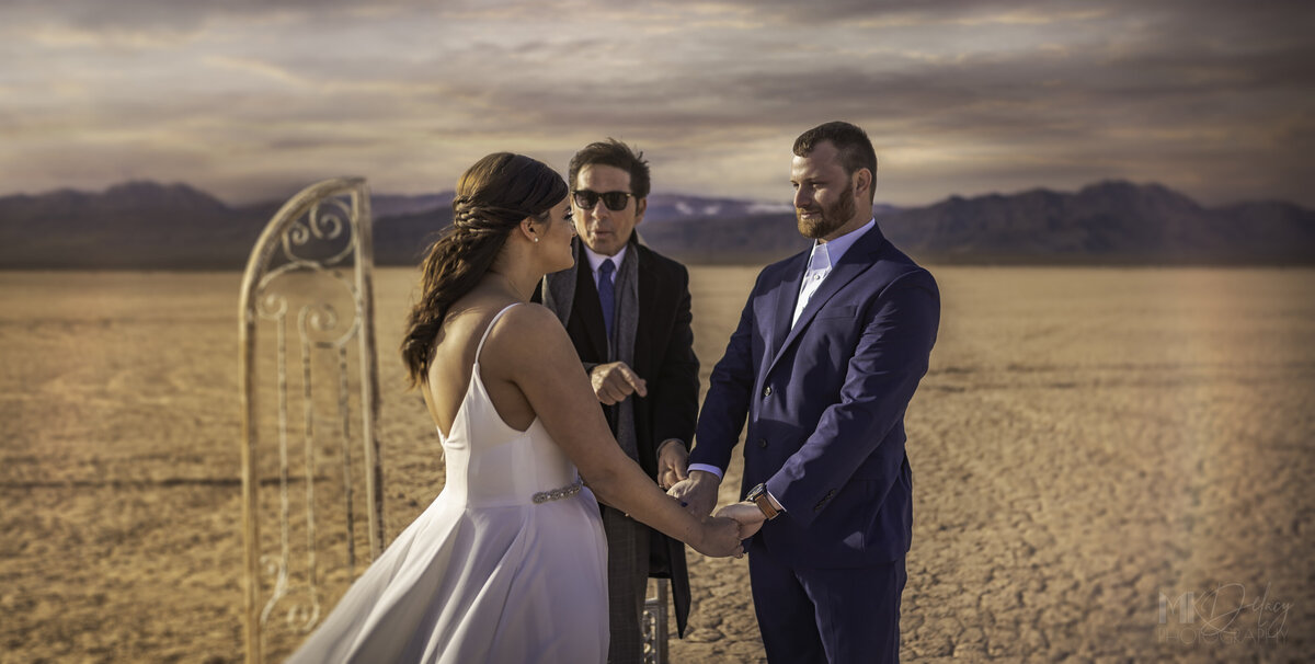 bride and groom holding hands and saying their vows in the desert with elvis impersonator Dry lake Bed elopement Blue Suit on Groom  flowers by michelle  bride in cream color wedding dress with deep  plunging  neckline mountain skyline  sunset las vegas wedding photographers mk delacy photography