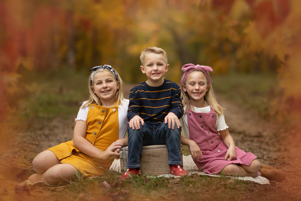 A boy and his 2 sisters sit for their Pella Family Portraits in the fall leaves