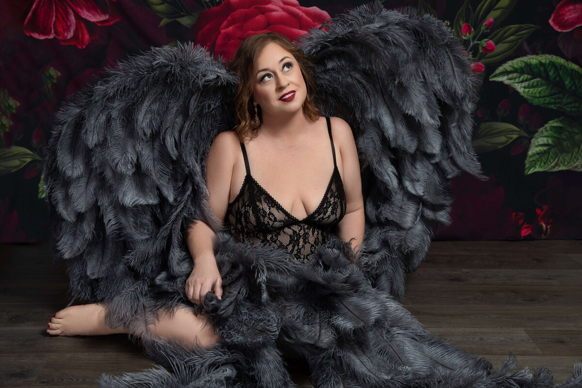 A woman with long dark hair wearing a black lace dress is seated on the floor against a richly colored floral backdrop. She has large, dark, feathery wings and is looking upwards with a thoughtful expression.