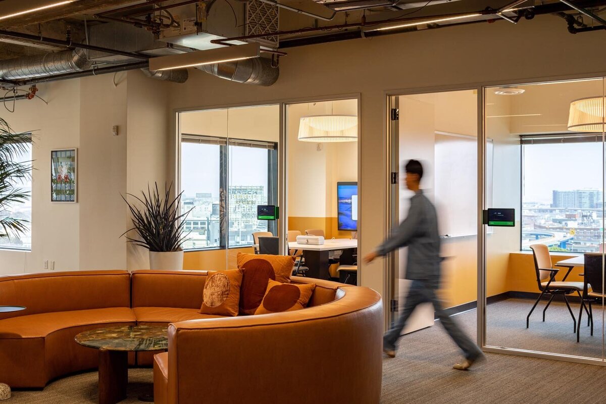 Collaborative office space with a curved leather sectional, glass-walled meeting rooms, and a person walking past in motion blur.