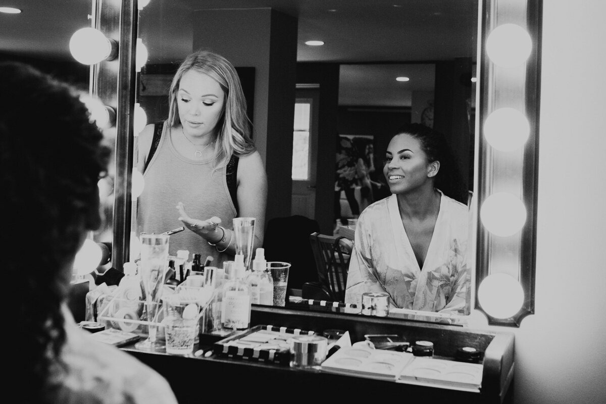 black and white image of a bride and make-up artist applying beauty products in the lighted mirrors at Willowbrook wedding venue preparation suite