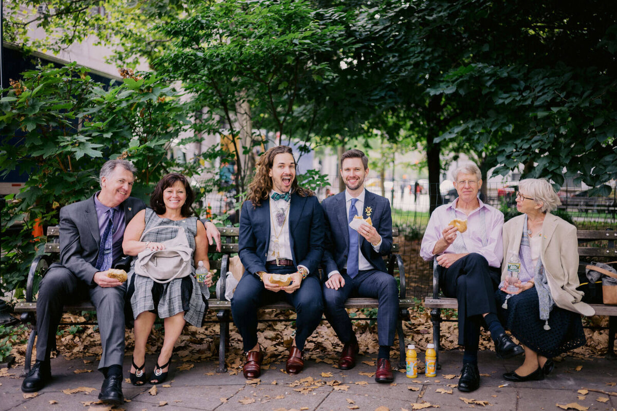 The two grooms and their parents are eating burger while sitting on a park bench. NYC City Hall Elopement Image by Jenny Fu Studio