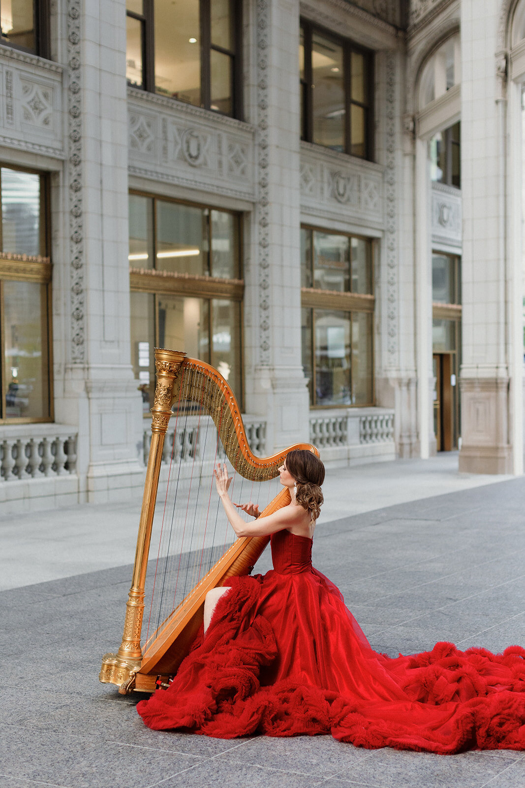 woman in long red gown draped behind her playing gold harp. She is seated in the urban courtyard at Wrigley Building in Chicago.