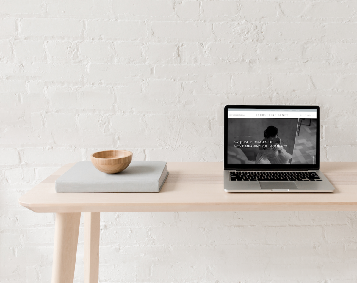 Macbook  displaying the image of a website on top of a light colored wooden table against a white brick wall.