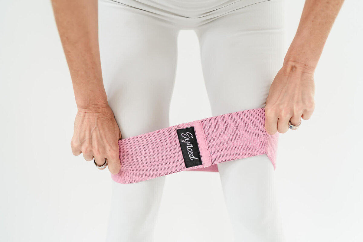Close-up of hands stretching pink Synced resistance band across white leggings.