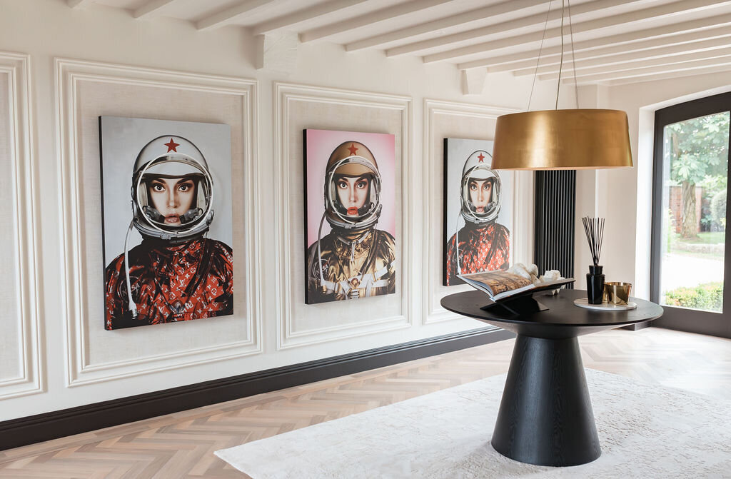 A modern interior features three portraits of astronauts with white helmets, framed on a white paneled wall. Centered is a black round table with decorative objects beneath a gold ceiling light.