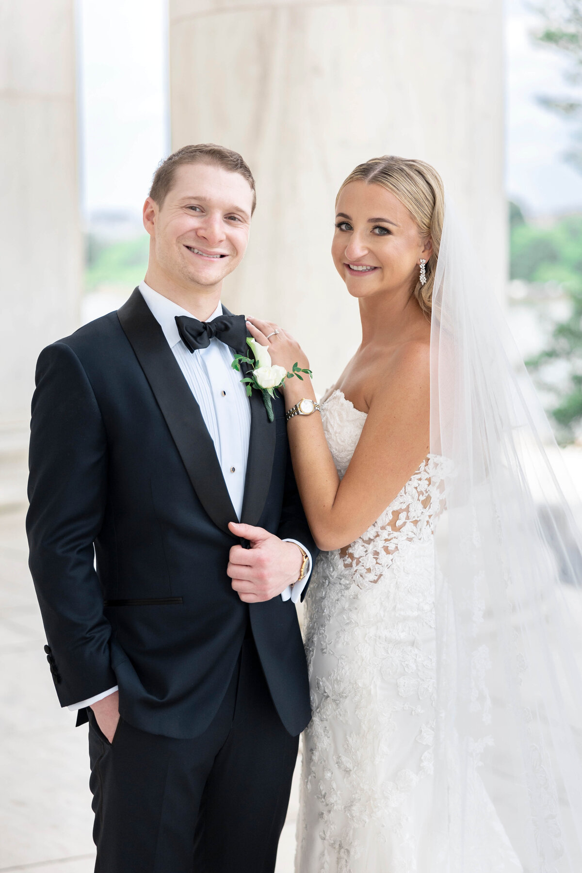 A bride and groom stand in front of a marble column. The bride is wearing a lace wedding dress and veil, smiling while gently holding the grooms arm. The groom is in a black tuxedo with a bow tie and white boutonniere. Both are smiling.