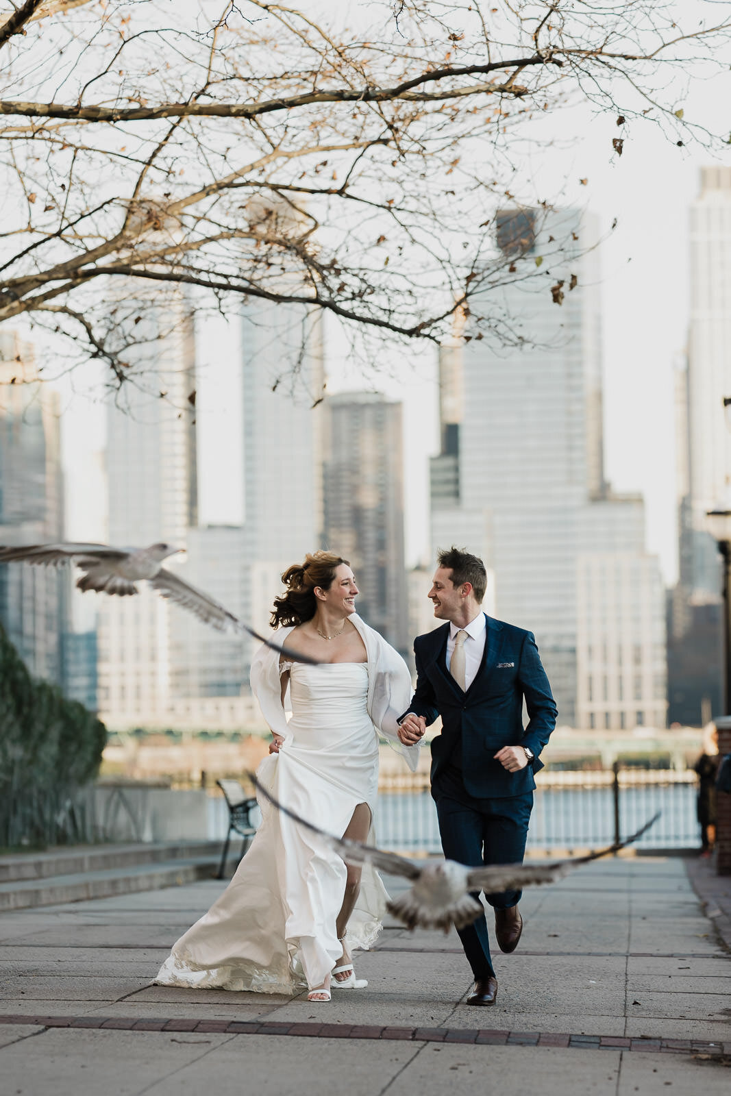 Bride and groom running hand in hand through Brooklyn Bridge Park with seagulls flying past and the New York City skyline in the background during a joyful candid wedding moment