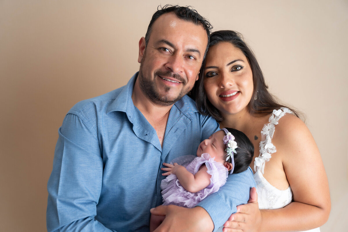 Close-up of a man cradling a newborn baby girl in his arms, with a woman leaning in closely, gently snuggling both.