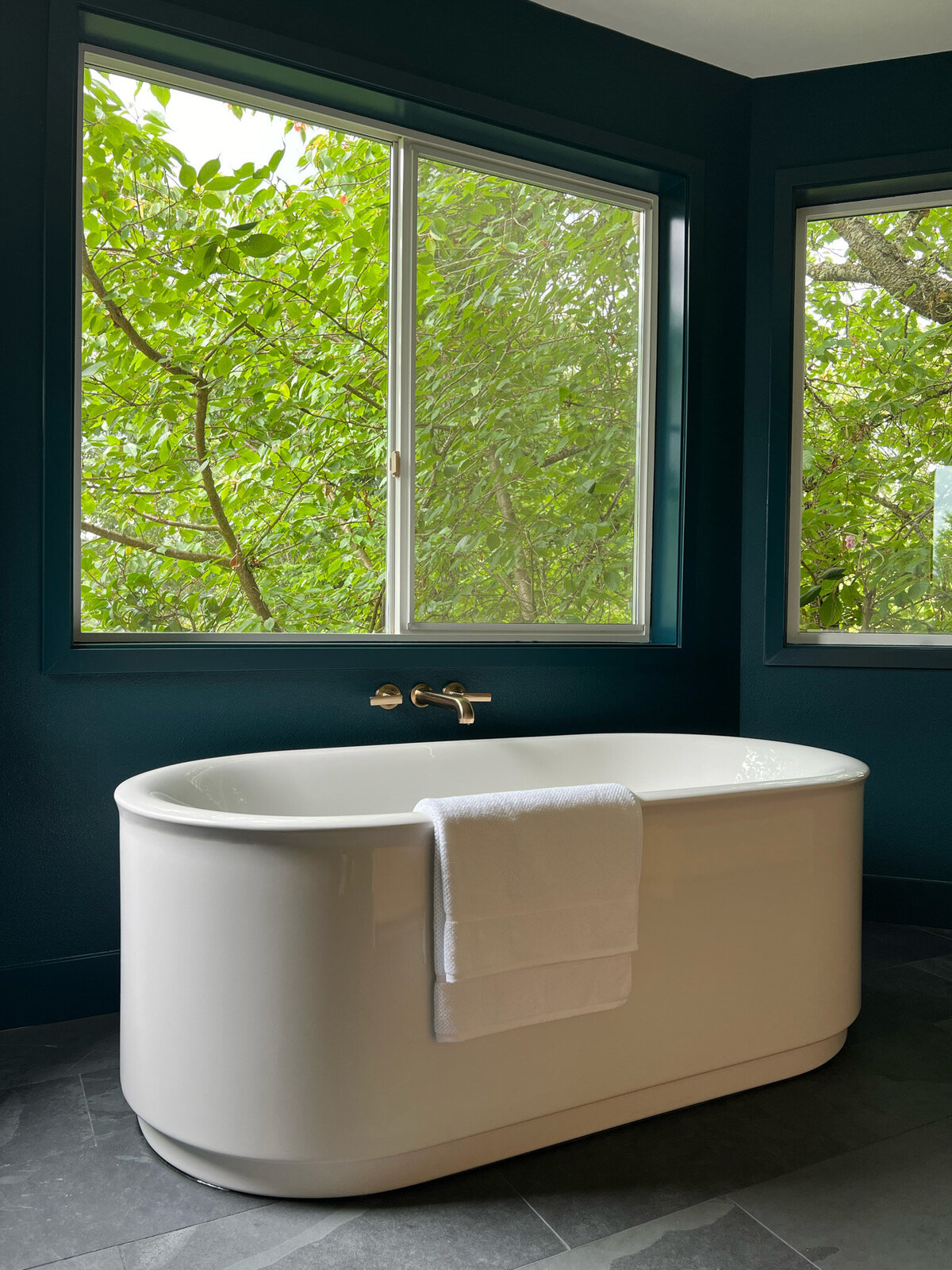 Bathroom with white freestanding bathtub in front of windows with a picturesque tree view. White towel draped over the bathtub. Dark teal blue walls.