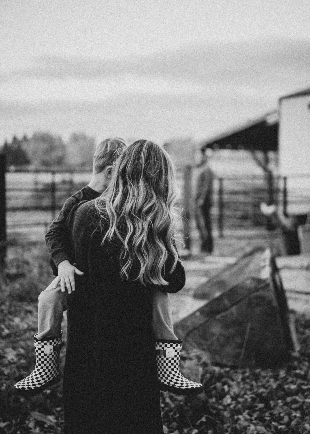 And indistinct, black and white image of a mother, carrying her young son, who is wearing black and white checkered rain boots on their family farm as they walk towards a barn to get their chores done.