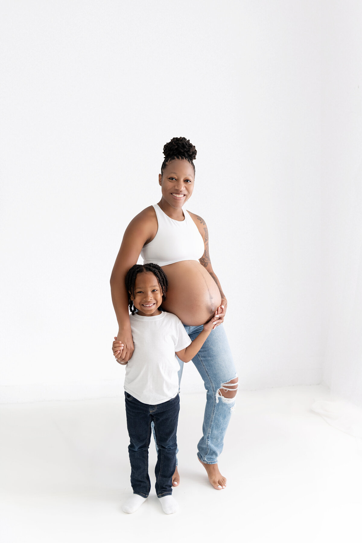 An expecting mother stands in a studio in jeans with her toddler son holding the bump with her