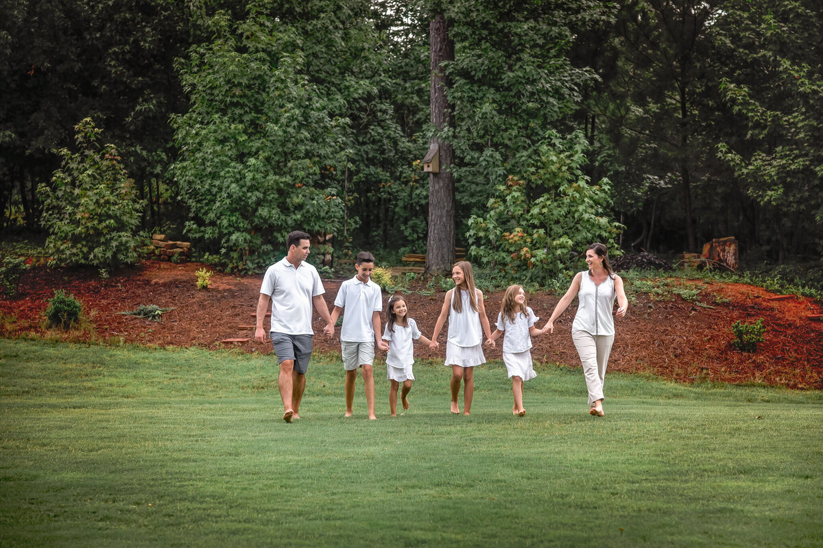 A family of 6 is dressed in white and holding hands while they walk outside during their family photo session.