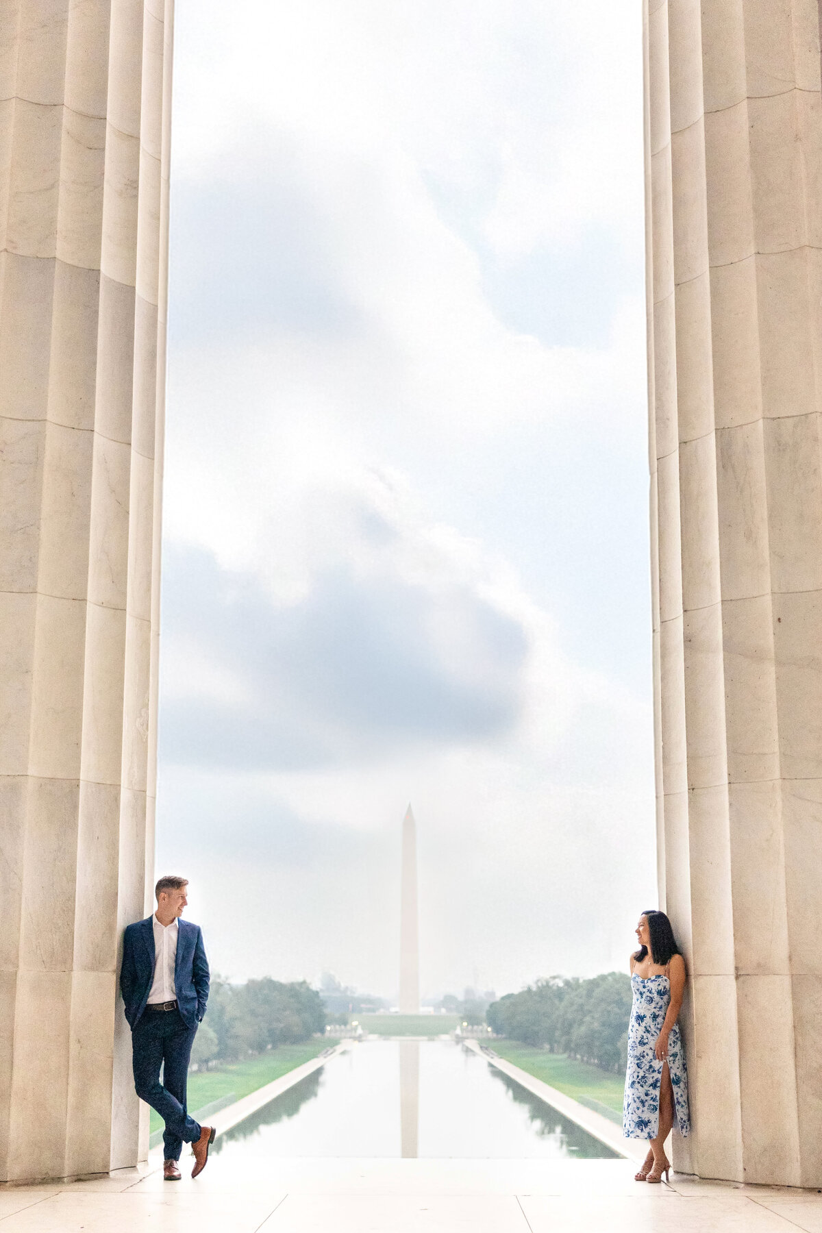 A man and woman stand apart, facing each other between the columns of the Lincoln Memorial. The Washington Monument is visible in the background, reflected in the Reflecting Pool under a partly cloudy sky.