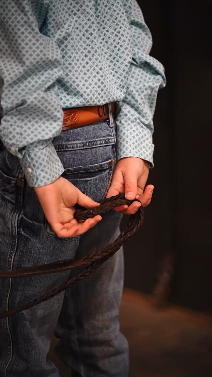 A small child wearing jeans and a blue western riding shirt, holds reins in his tiny hands