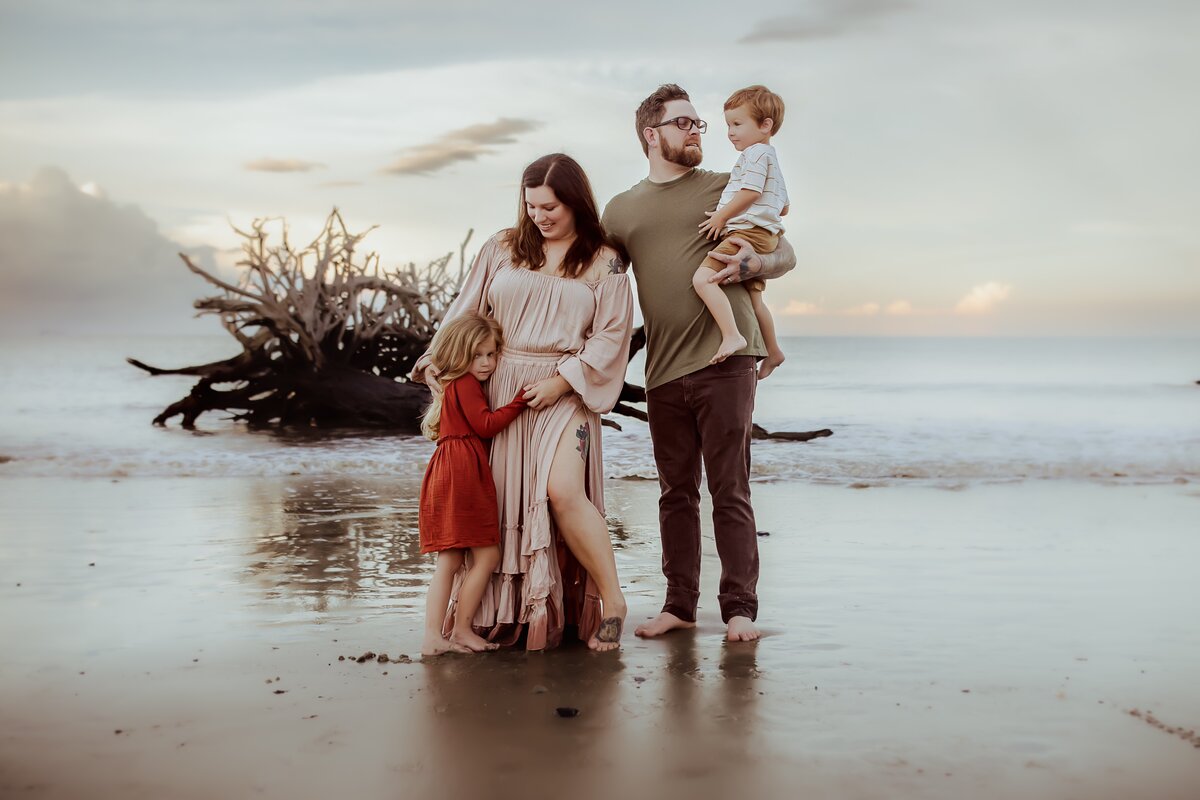 A family of four stands on a driftwood beach. A woman holds a young girl in a red dress, while a man carries a small child wearing a white outfit. The sea and cloudy sky serve as the backdrop, capturing the essence of family photography amidst nature's beauty.