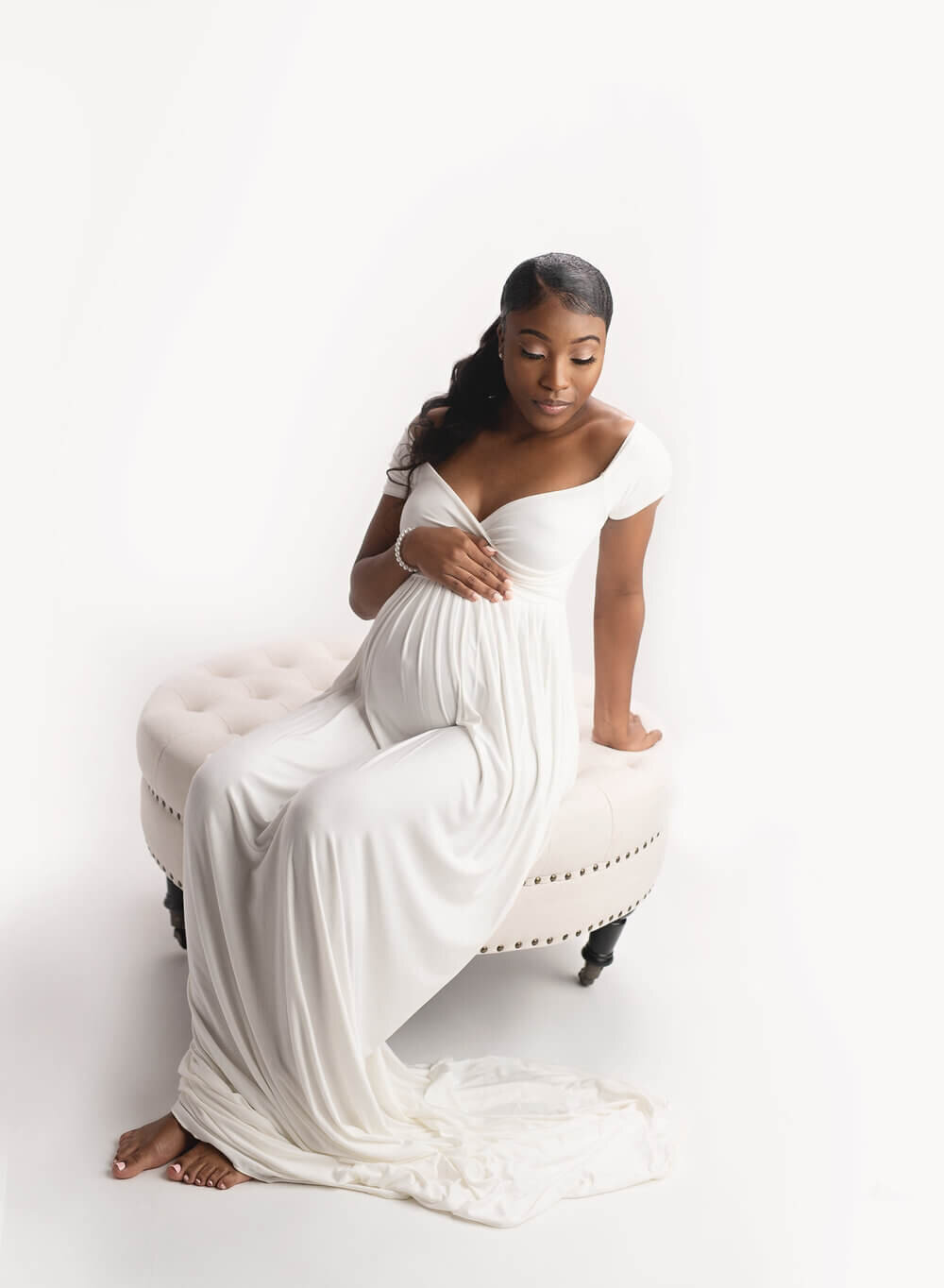 A mother to be sits on a white stool in a studio wearing a white maternity gown