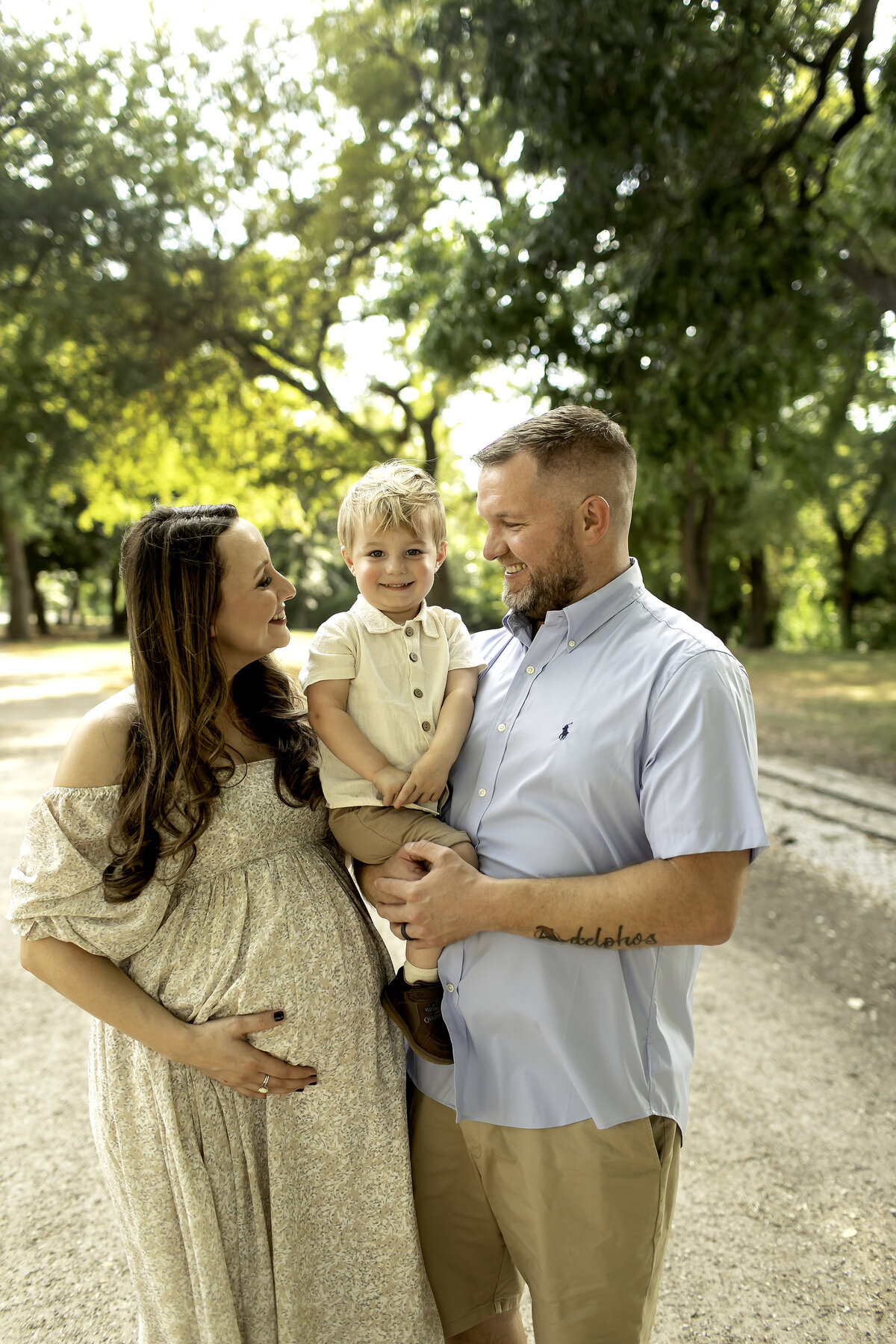 A joyful maternity photo of mom, dad, and their big brother smiling together in Trinity Park, with the lush greenery and scenic paths in the background, captured by Chunky Monkey Photography.