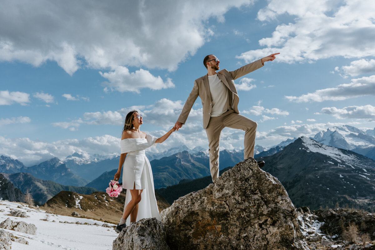 Groom pointing towards mountains while standing on rock with bride