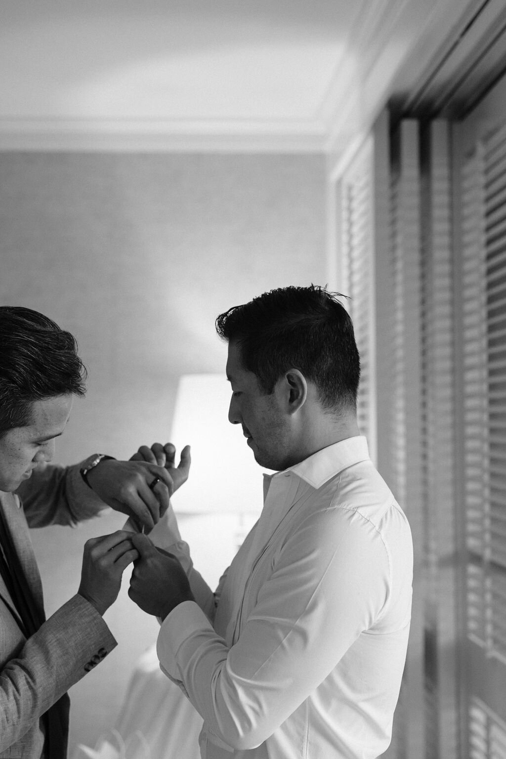 black and white of groom getting cufflink put on in hotel room by groomsman
