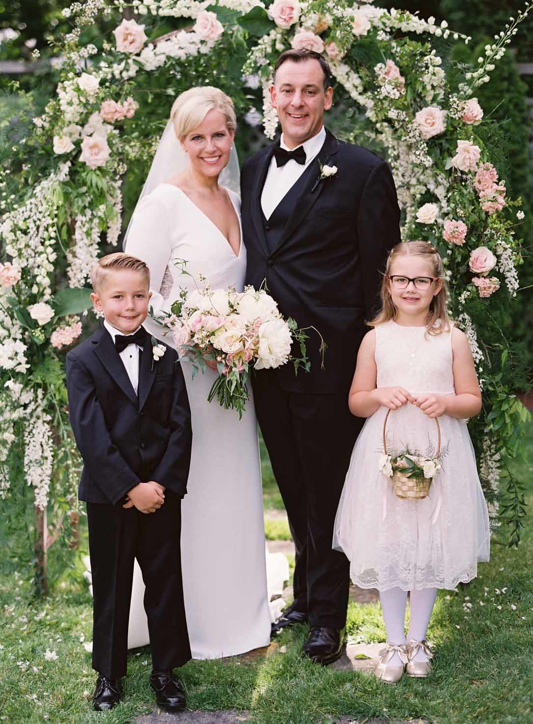 bride and groom with flower girl and ring bearer in front of flower covered wedding arbor