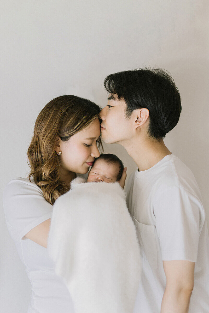 Mother is holding her newborn baby in her arms and the father is kissing on the mother's forehead. A simple yet beautiful photo of a newborn session in Yokohama.