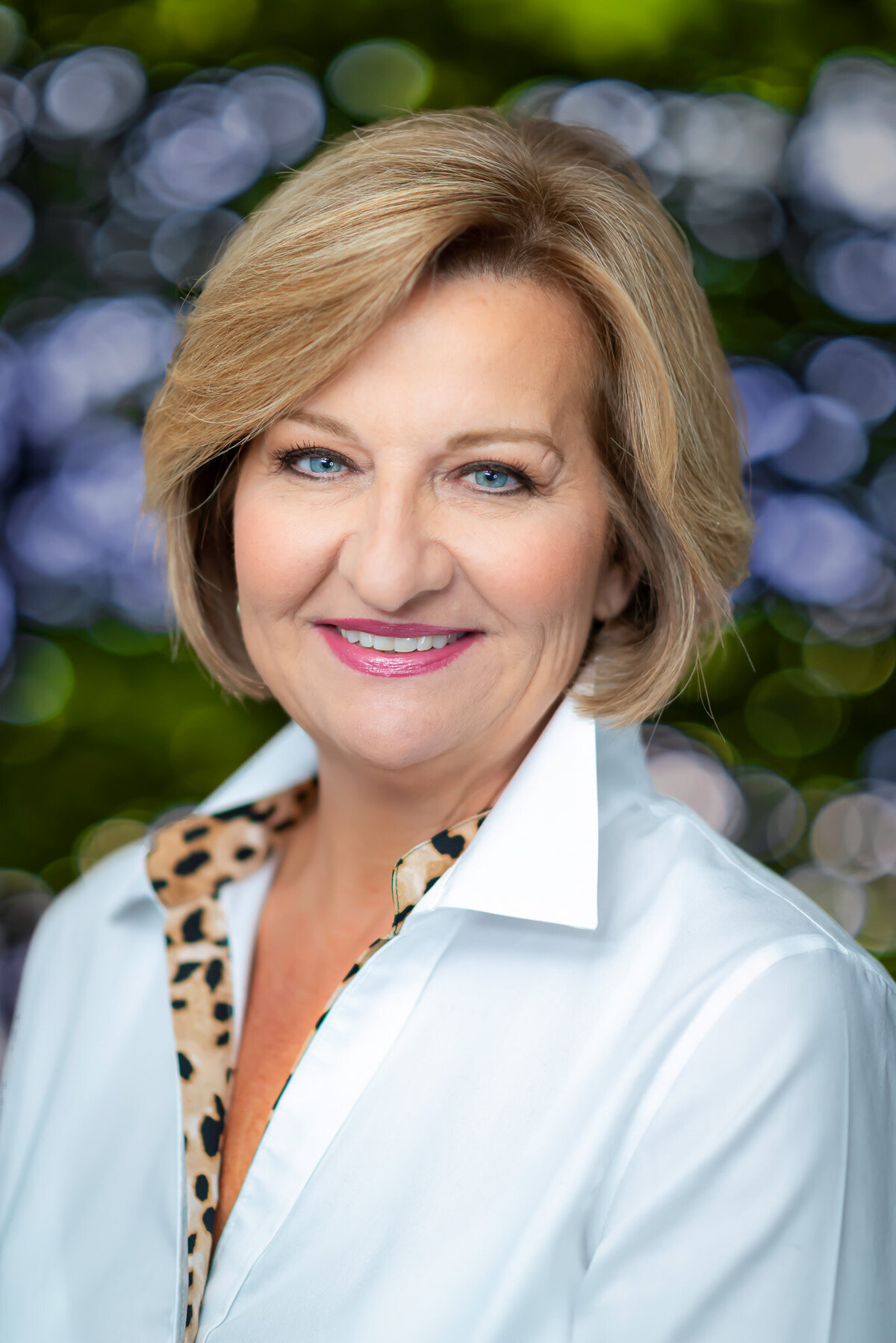 A woman in a white shirt poses in front of a bush with purple flowers for a headshot.