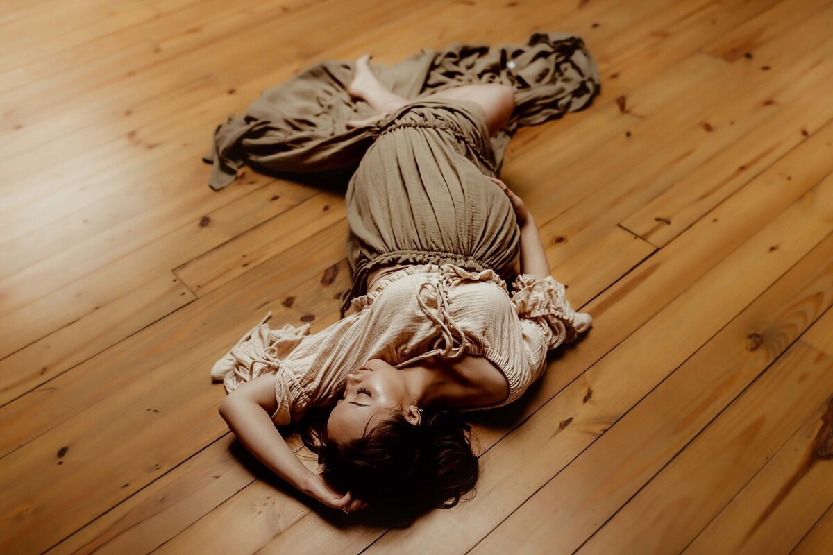 A woman in a beige dress lies on a wooden floor, eyes closed, with an arm bent over her head.