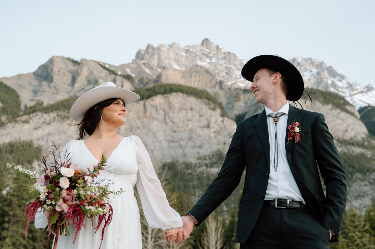 Bride and groom in western attire walk hand in hand with towering mountains in the background in Banff.