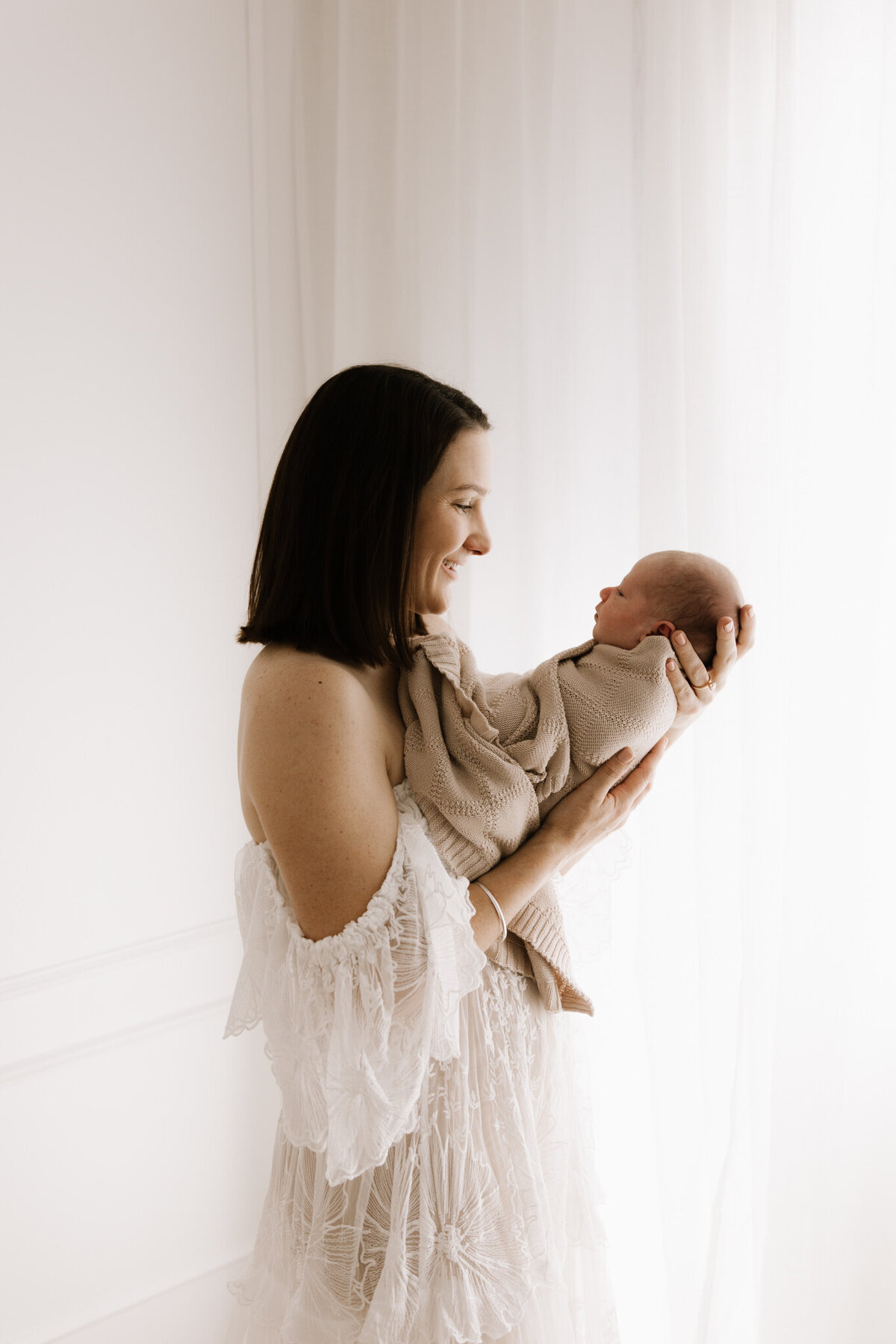 Smiling mother in a lace dress gently cradling her newborn baby by the window, captured in soft studio lighting by a newborn photographer in Southwest Sydney.