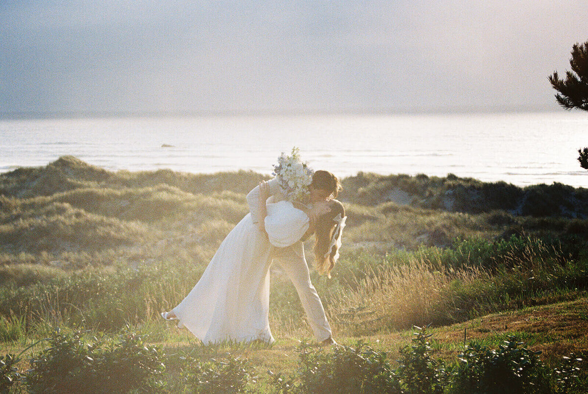 A groom bends his bride and kisses her with the ocean in the background.