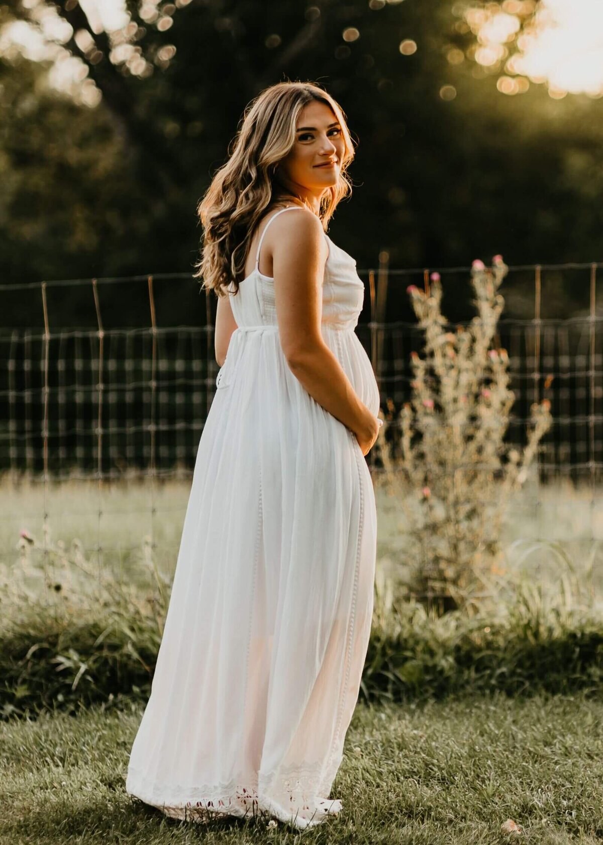A pregnant woman in a white dress stands elegantly in a field, captured by a Pittsburgh maternity photographer.