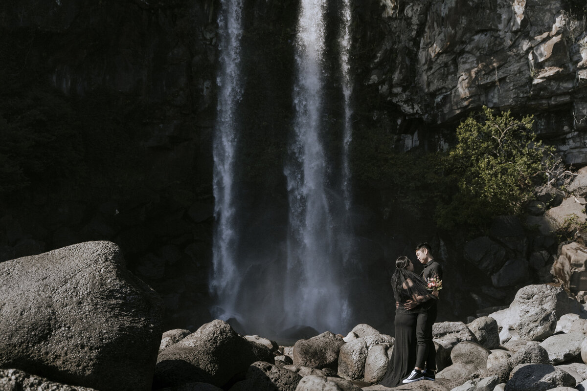 bride wearing a  black long dress and the groom in his black suit at Jeongbang Waterfall in Jeju
