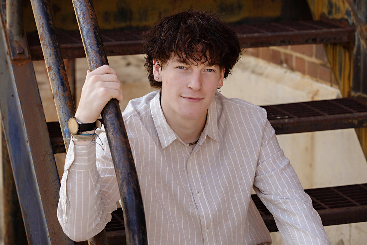 High school senior sitting on a rusted staircase at Camp North End in Charlotte NC, wearing a vertical stripe button down shirt and cream jeans.