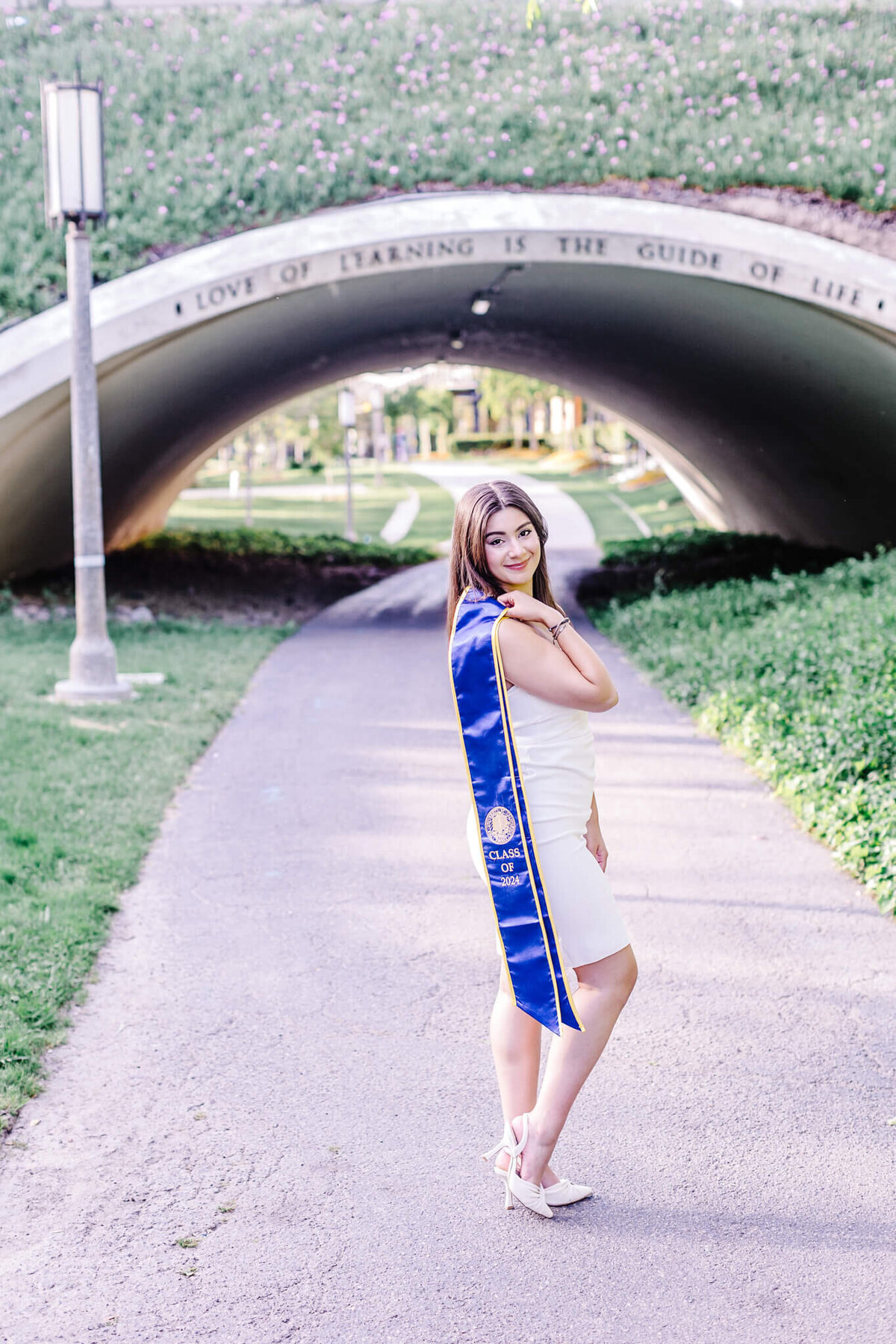 A woman in a white dress and blue graduation stole stands on a path beneath an archway with the words Love of Learning is the Guide of Life. She is looking over her shoulder, surrounded by greenery and soft daylight.