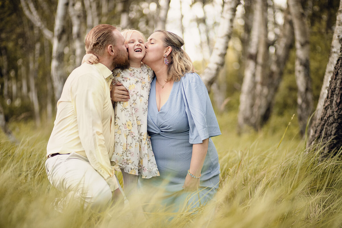 mamma och pappa som pussar sin dotter på varsin kind i högt gräs i börjskog