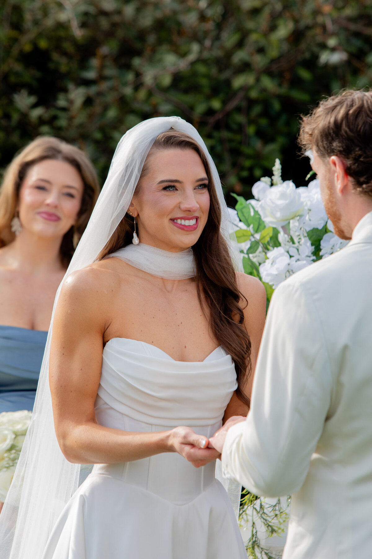 A bride smiles at her groom and holds his hands during their wedding ceremony. Behind her is one of her bridesmaids, admiring the couple.