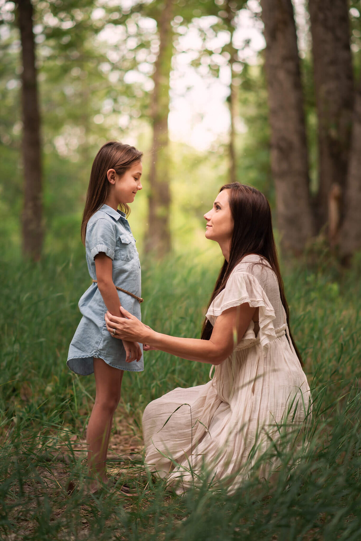 A mother kneels down in the tall grass while holding her duaghters hand in front of her. They look at eachother lovingly. Portrait by Brittany Danielle