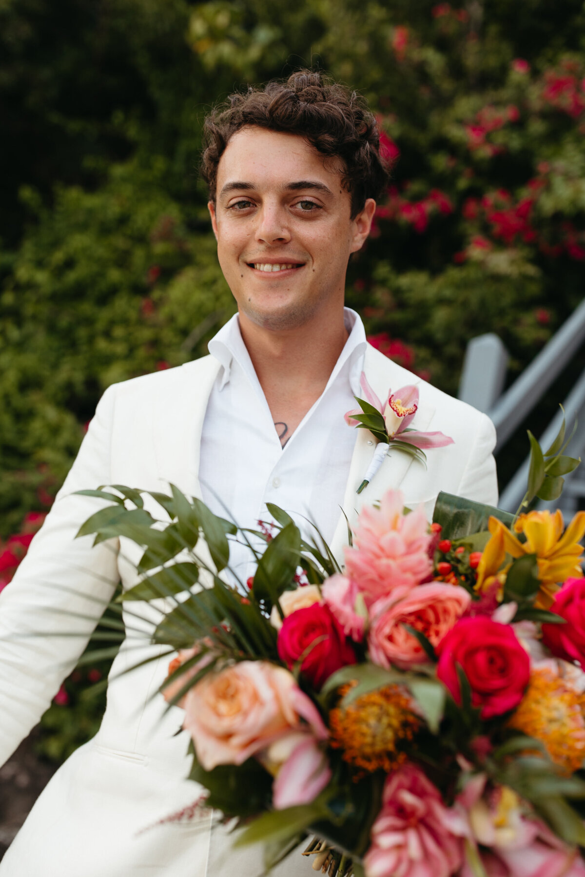 Groom holding a vibrant wedding bouquet, smiling warmly, with his white suit contrasting against the colorful flowers