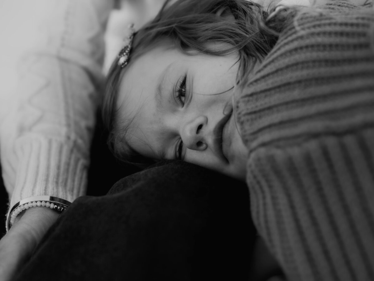 A close-up black-and-white portrait of an adorable, curly haired toddler laying on her mothers lap during it outdoor photo session.