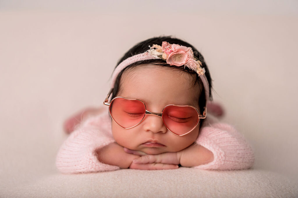 Adorable newborn baby girl wearing heart-shaped sunglasses and pink floral headband photographed by Connecticut newborn photographer in studio