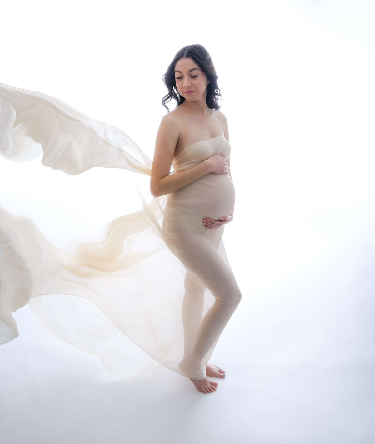 Mother-to-be in a side profile, looking over her shoulder, with flowing fabric tossed by a fan. Image captured by E. Benecki Photography.