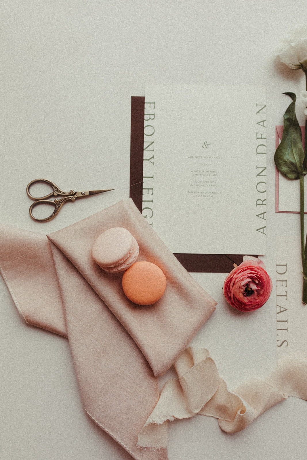 White wedding stationery with gray font set with a blush napkin, peach-colored macaroons, a flower and scissors.
