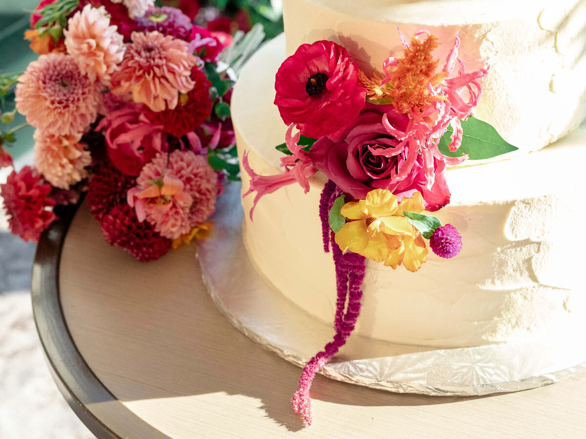 A two-tiered white cake adorned with vibrant flowers including red roses, pink blooms, and yellow accents. A matching floral arrangement is placed nearby on a table, creating a colorful and festive setting.