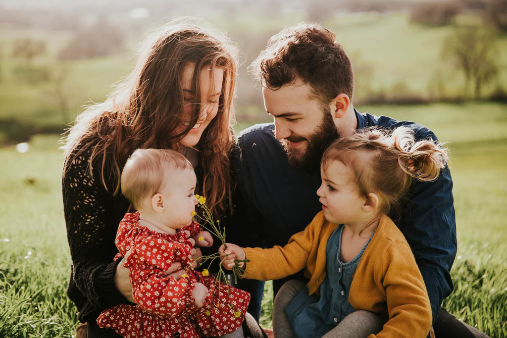 Family portrait sitting in grassy field with toddler offering a flower toward baby's face